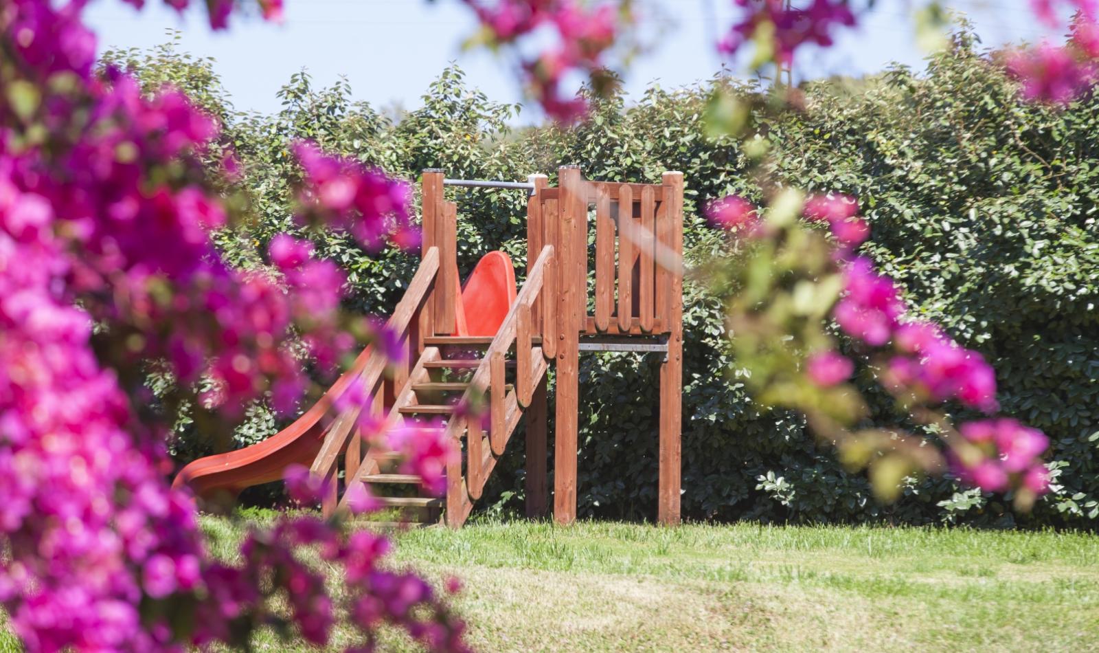 Scivolo in legno nel giardino, circondato da fiori rosa e verde.