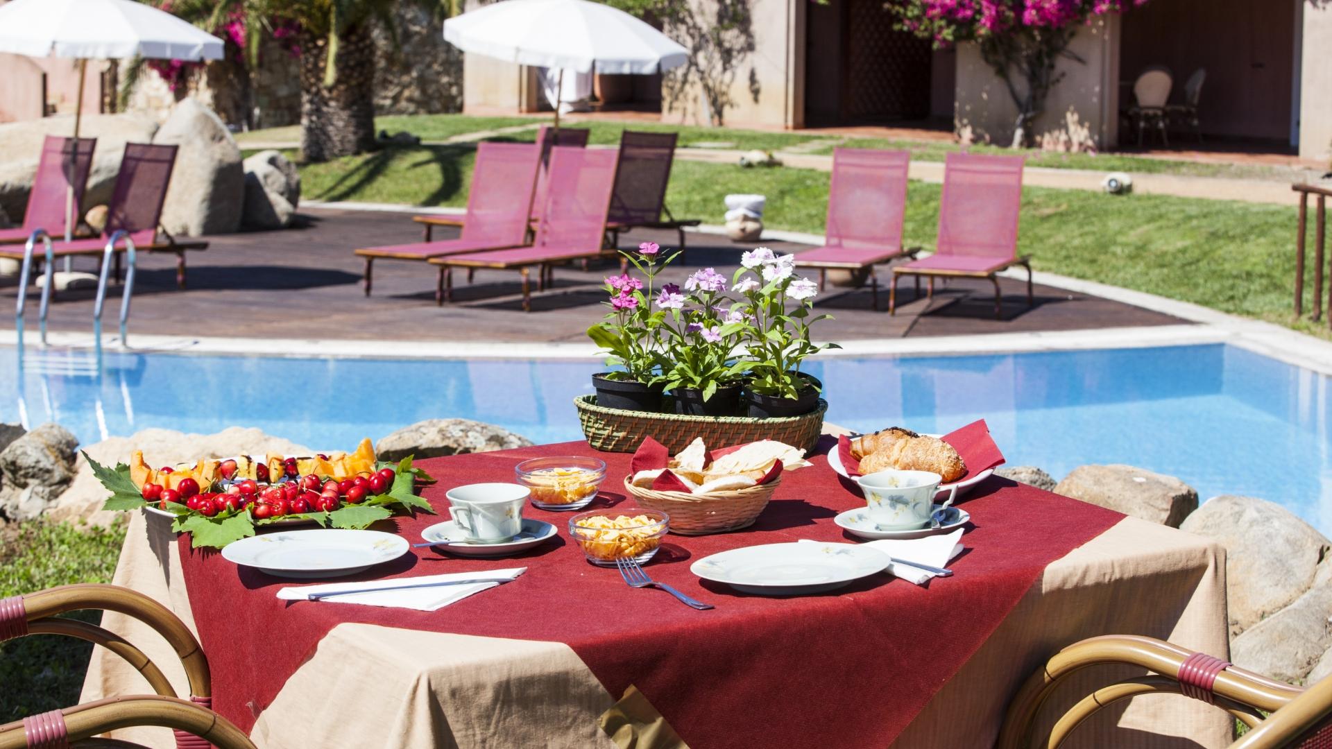 Colazione all'aperto vicino alla piscina, con frutta e fiori su tavolo apparecchiato.