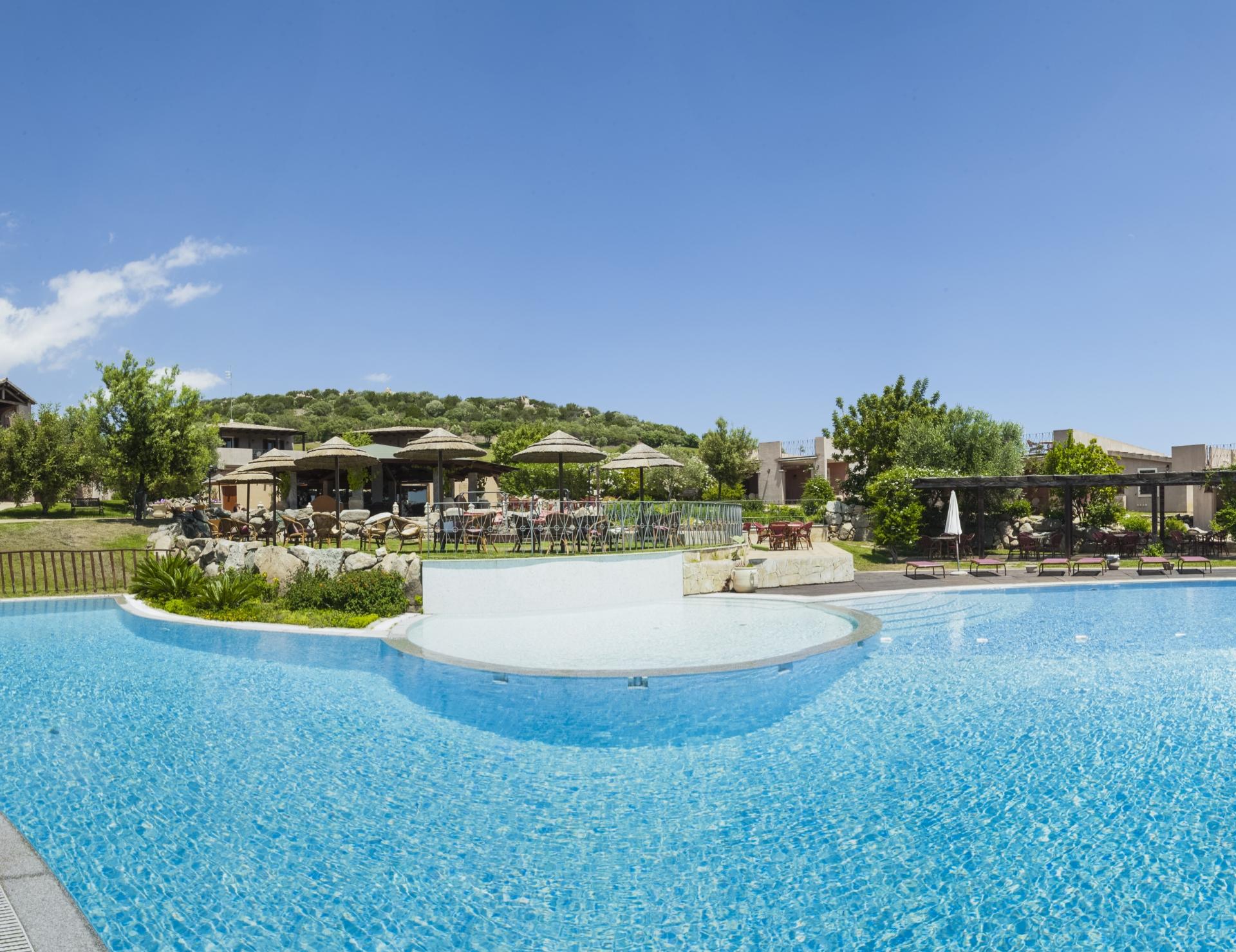 Piscina all'aperto con ombrelloni e giardino, sotto un cielo sereno.