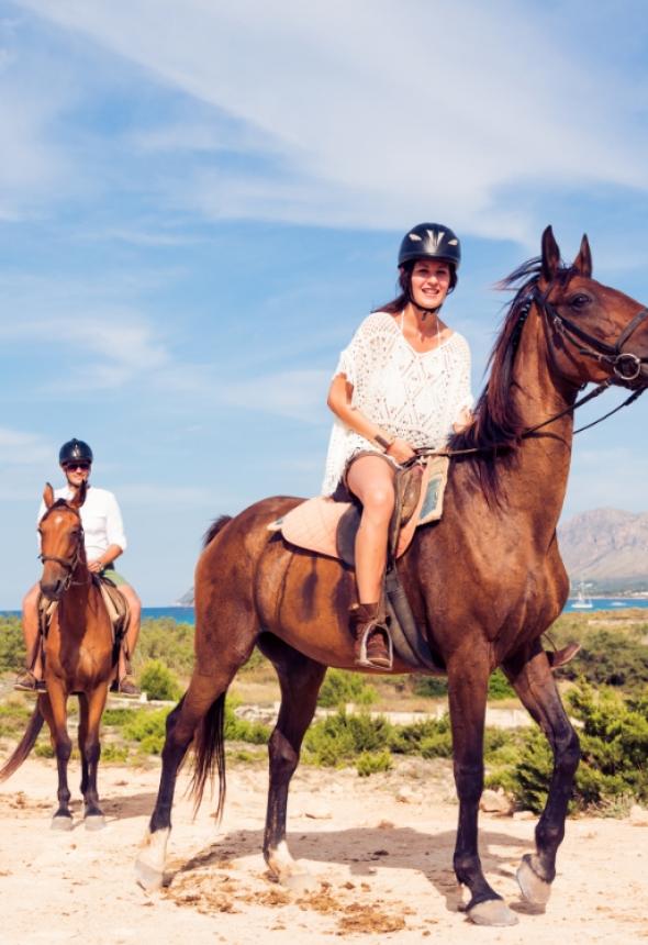 Due persone a cavallo su un sentiero panoramico, indossano caschi protettivi.